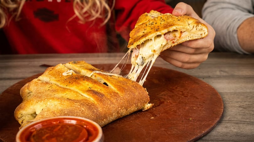 A hand pulls a slice of cheesy calzone, stretching melted cheese, from a whole calzone on a wooden board.