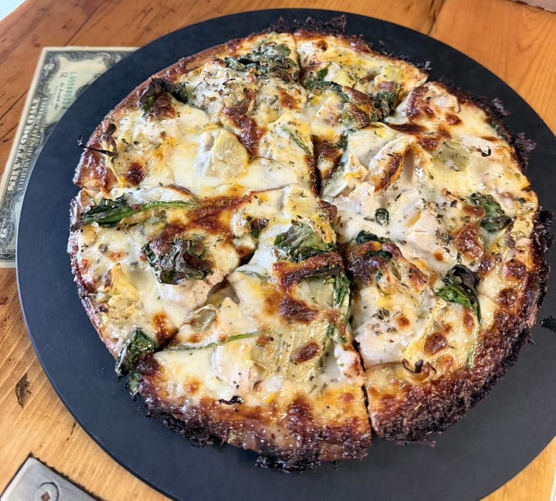 White pizza with spinach, artichoke, and chicken on a black plate, next to a two-dollar bill.