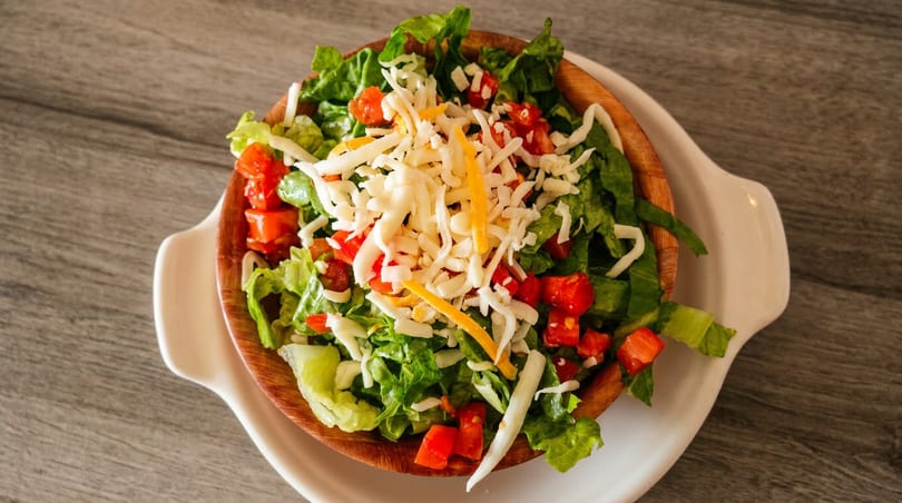 Fresh green salad with diced tomatoes and shredded cheese in a wooden bowl on a white plate.