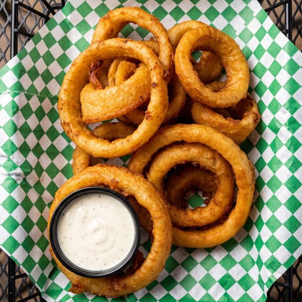 Golden-brown onion rings and a small cup of ranch dressing in a black wire basket with green and white checkered paper.