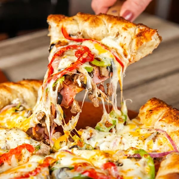 Hand lifting a slice of loaded pizza with melted cheese stretching, on a wooden table.
