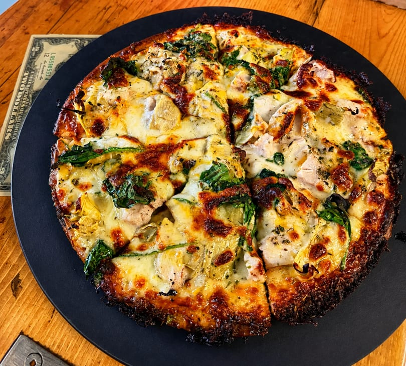 A sliced pizza with melted cheese, spinach, artichoke hearts, and chicken on a black plate.
