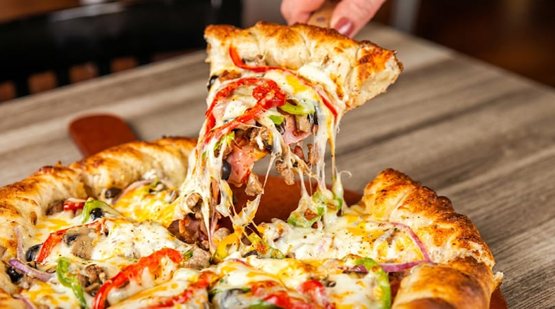 A hand lifts a slice of loaded pizza with melted cheese stretching from the pie.